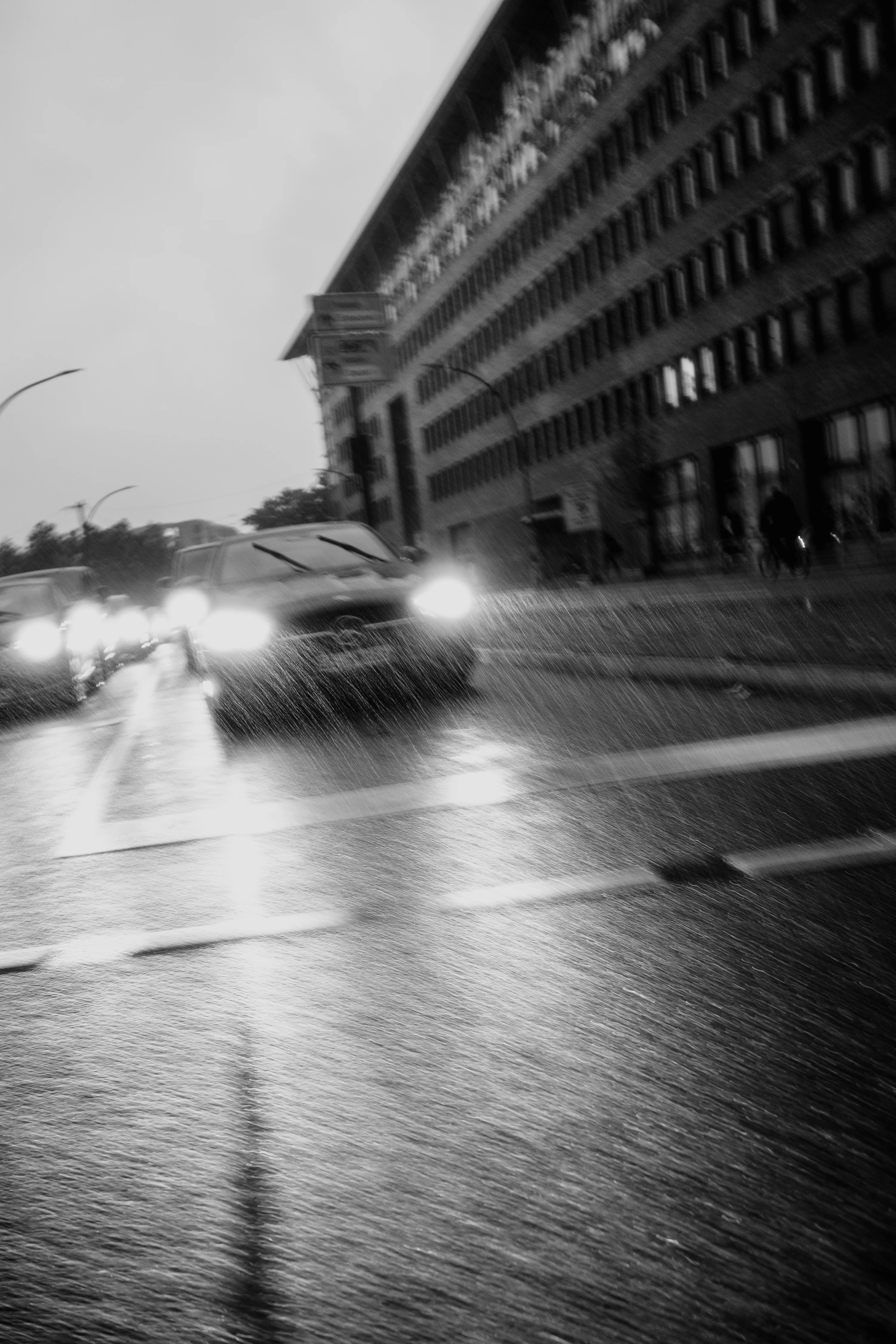 Black and white photo of cars on a rainy street with blurred headlights in front of a tall building.