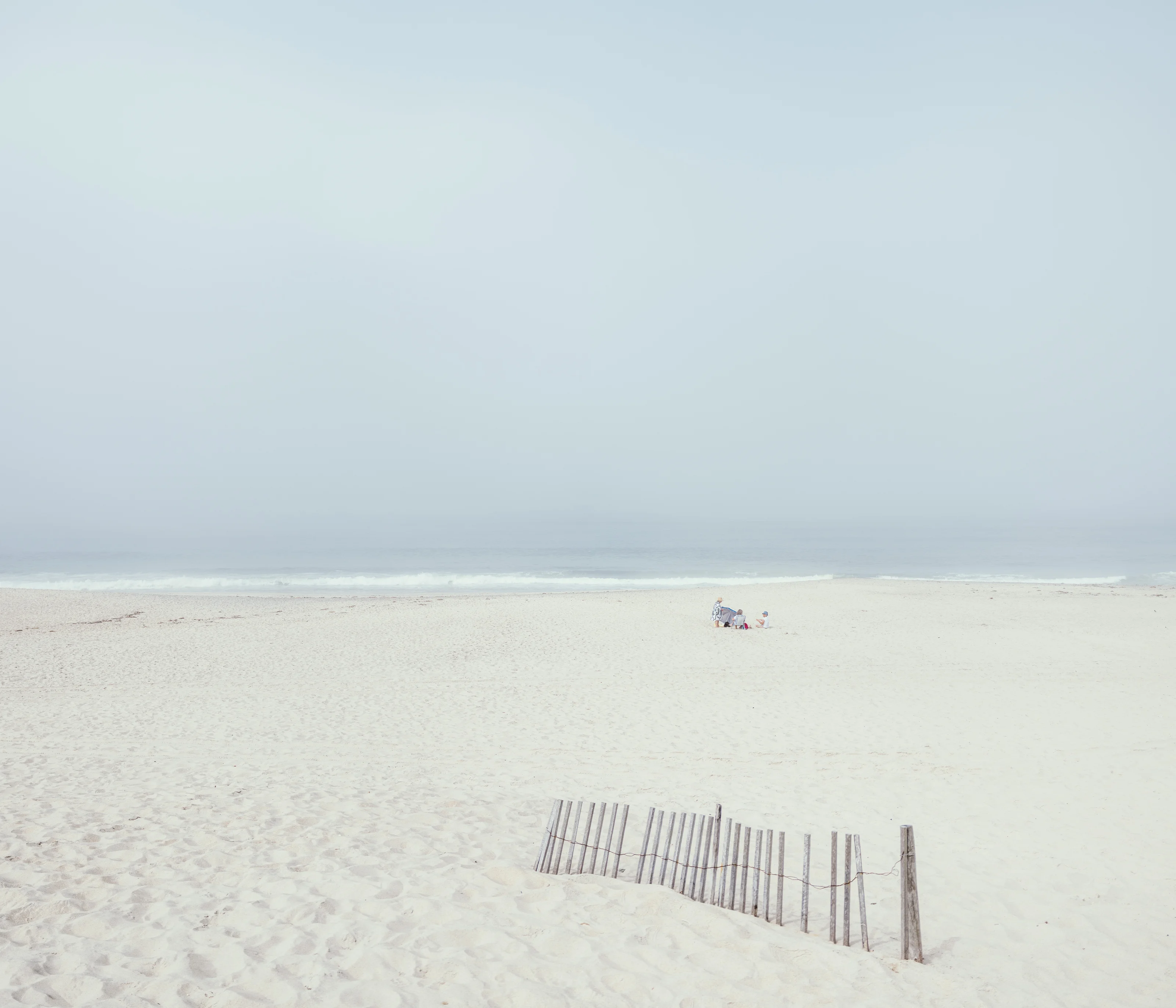 A sparse beach scene with a thin, uneven wooden fence in the foreground and a small group of people sitting near the water's edge.