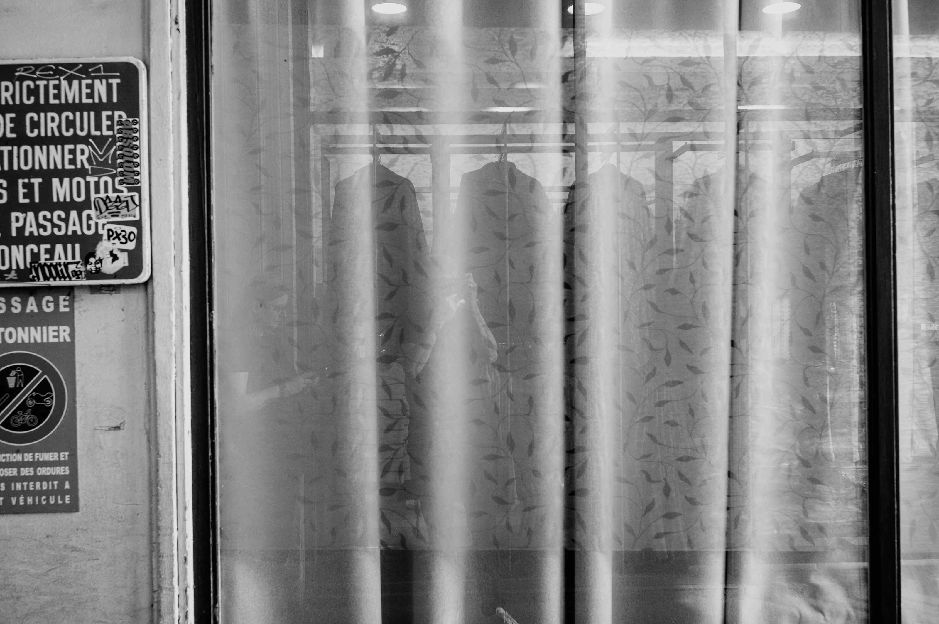 Black and white photo of a patterned curtain with shadows of hanging clothes behind, and partially obscured urban signs on the left.