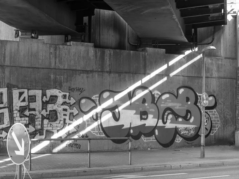 Graffiti on a concrete wall under a bridge with sunlight streaming in diagonally.