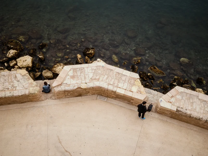 Two people by a stone wall overlooking a rocky shoreline with a wide expanse of water.