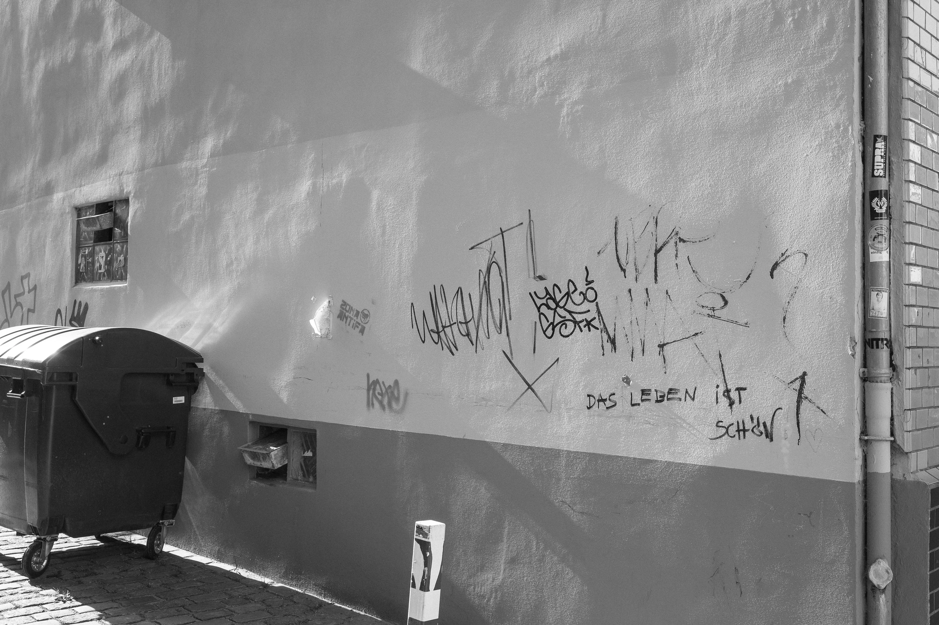 A wall with various graffiti tags and a garbage bin on a cobblestone street.