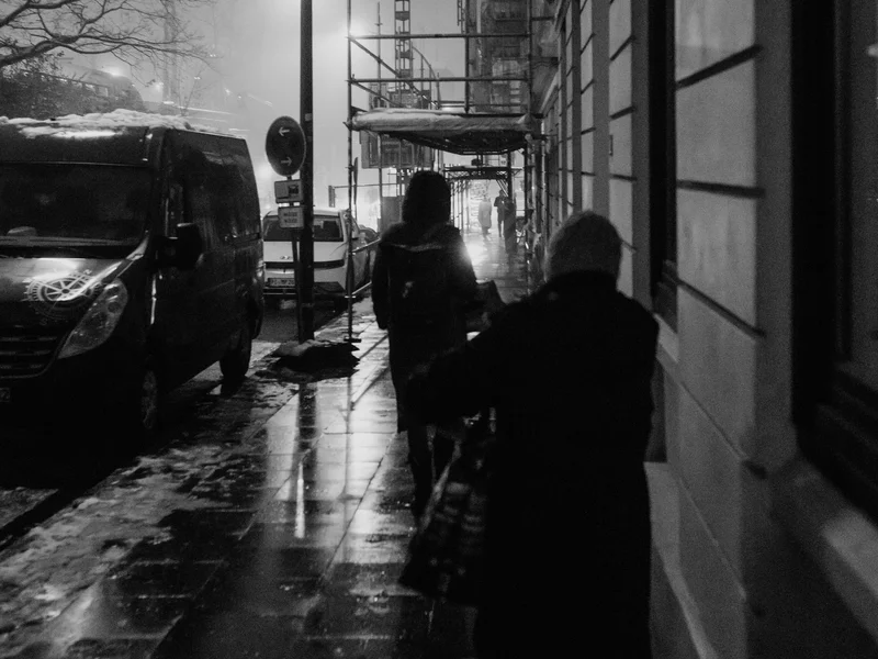 Two people walking on a wet, dimly lit street at night with buildings and cars lining the side.