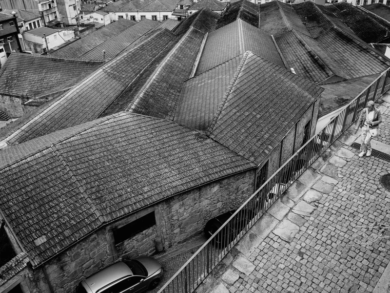 Black and white photo of tiled rooftops descending towards a river, with a person walking on a cobblestone path.