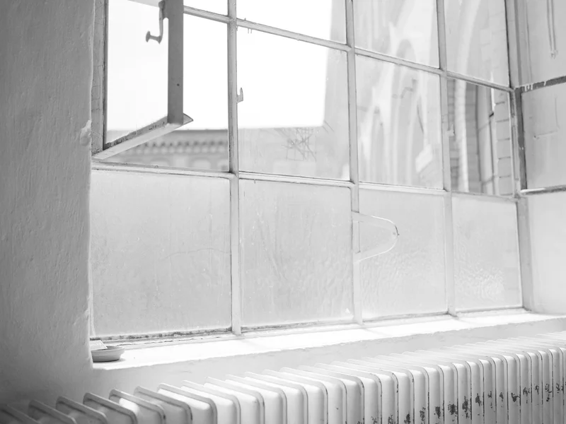 A black and white photo of a radiator beneath a large, multi-paned window with an open section.
