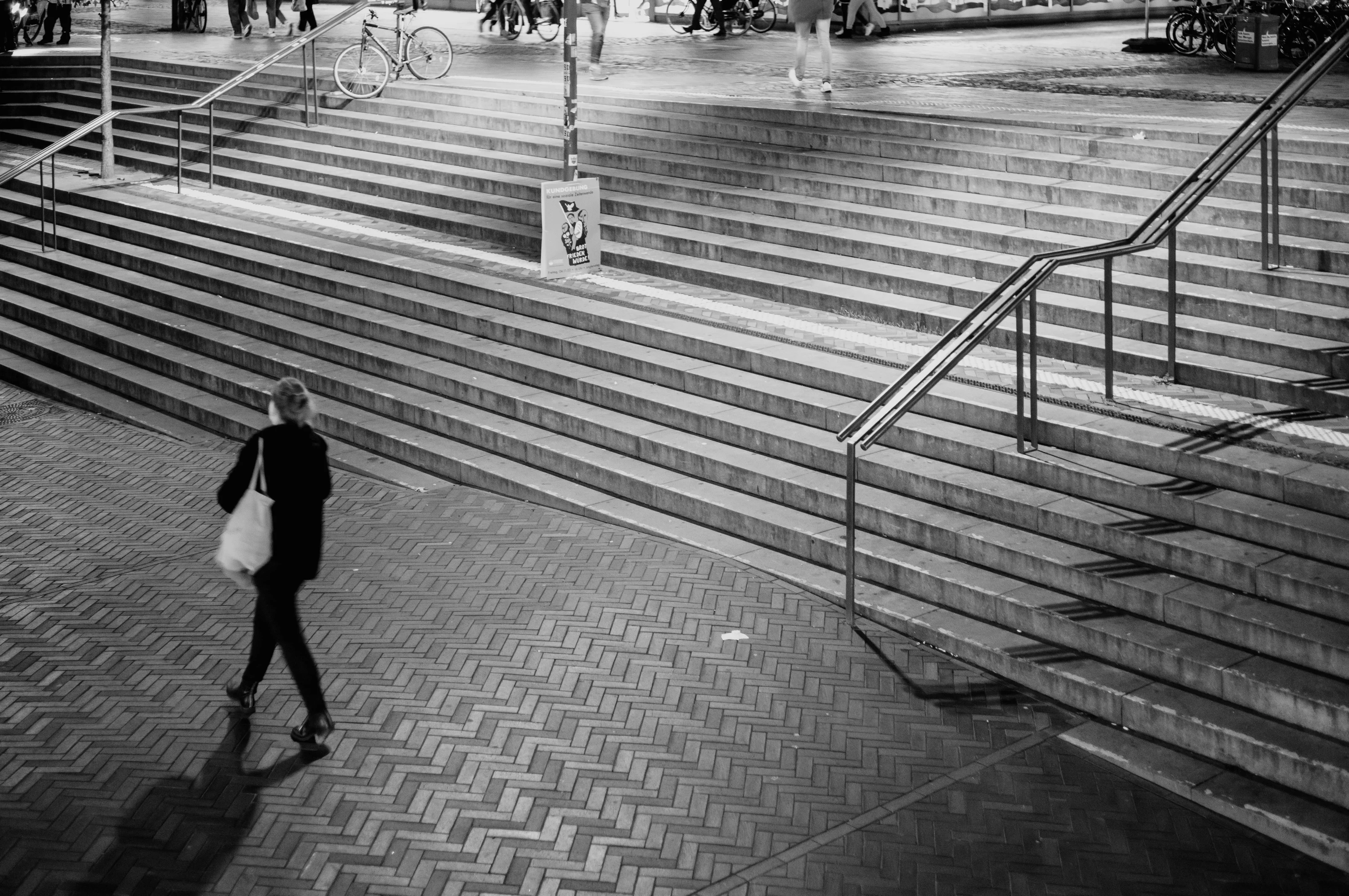 A person walks across a herringbone brick pattern with stairs and railings in the background.