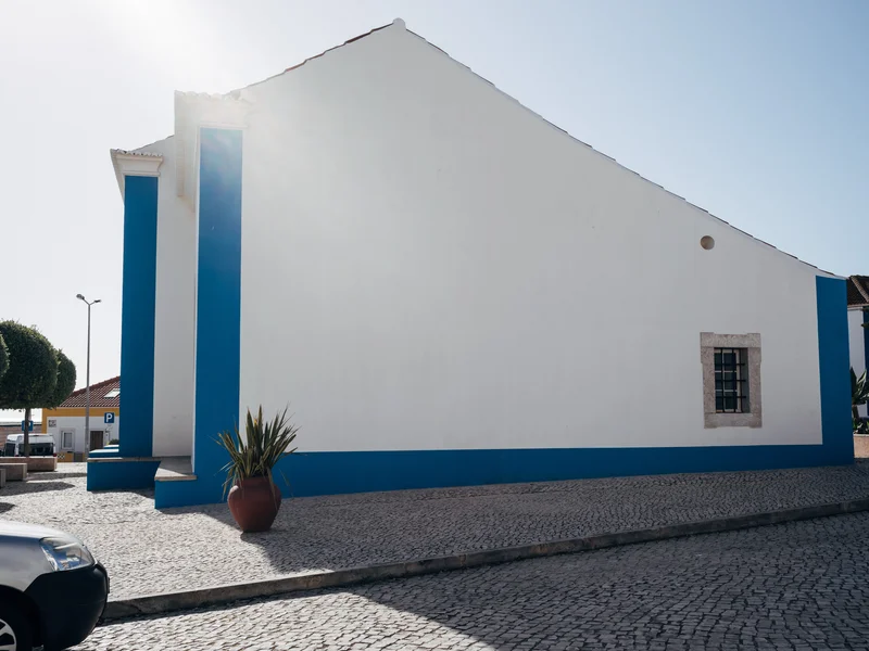 White building with blue accents under a clear sky, with sunlight creating shadows on the cobblestone ground.