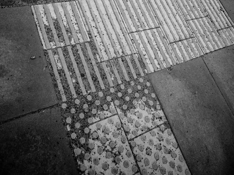 Black and white photo of a textured sidewalk edge with tactile paving next to a road with dashed lines.