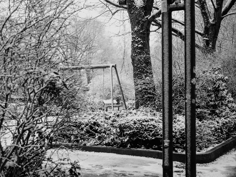 Snow-covered playground with a swing surrounded by trees and bushes.