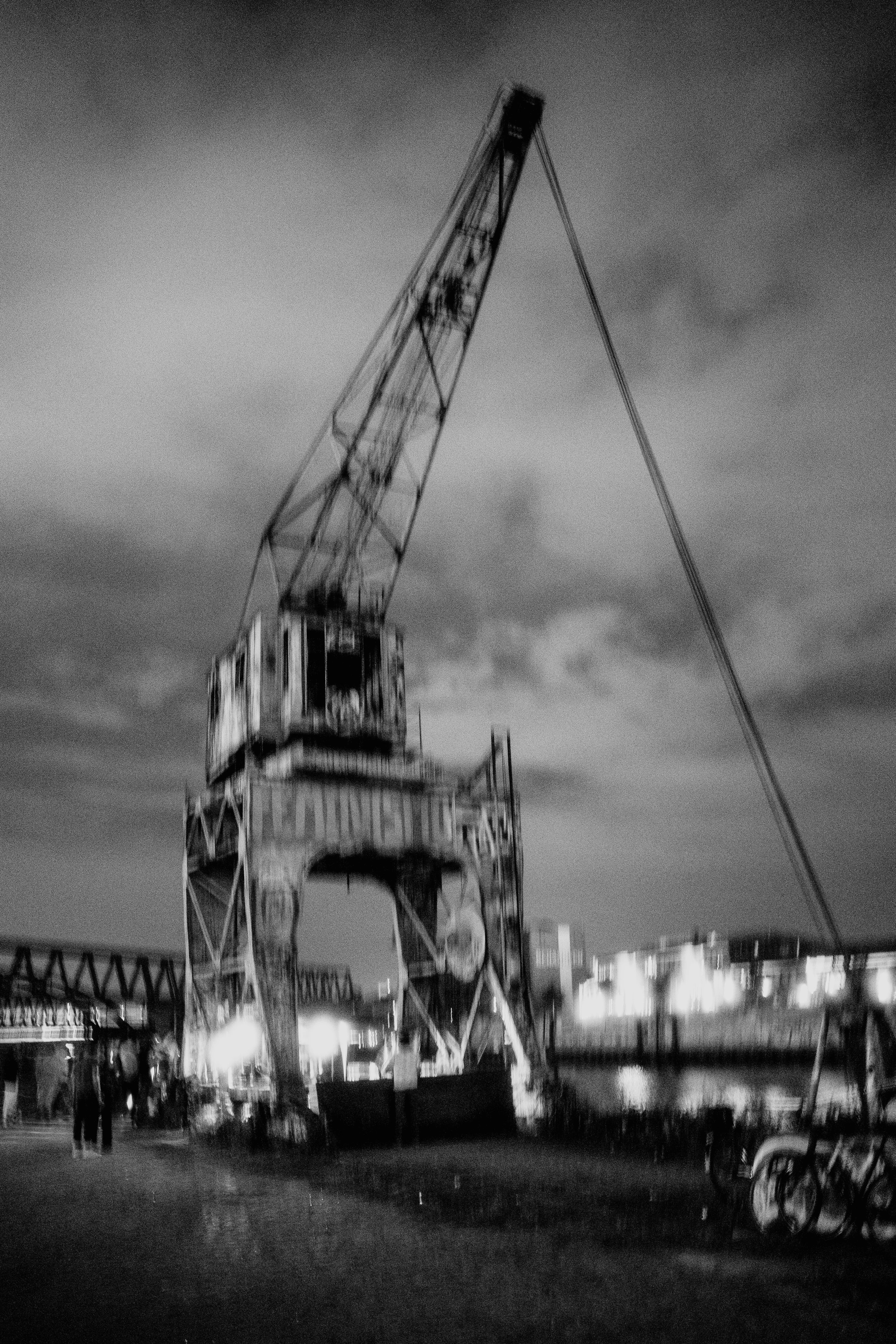 Blurry black and white image of an industrial crane by a waterfront at night.