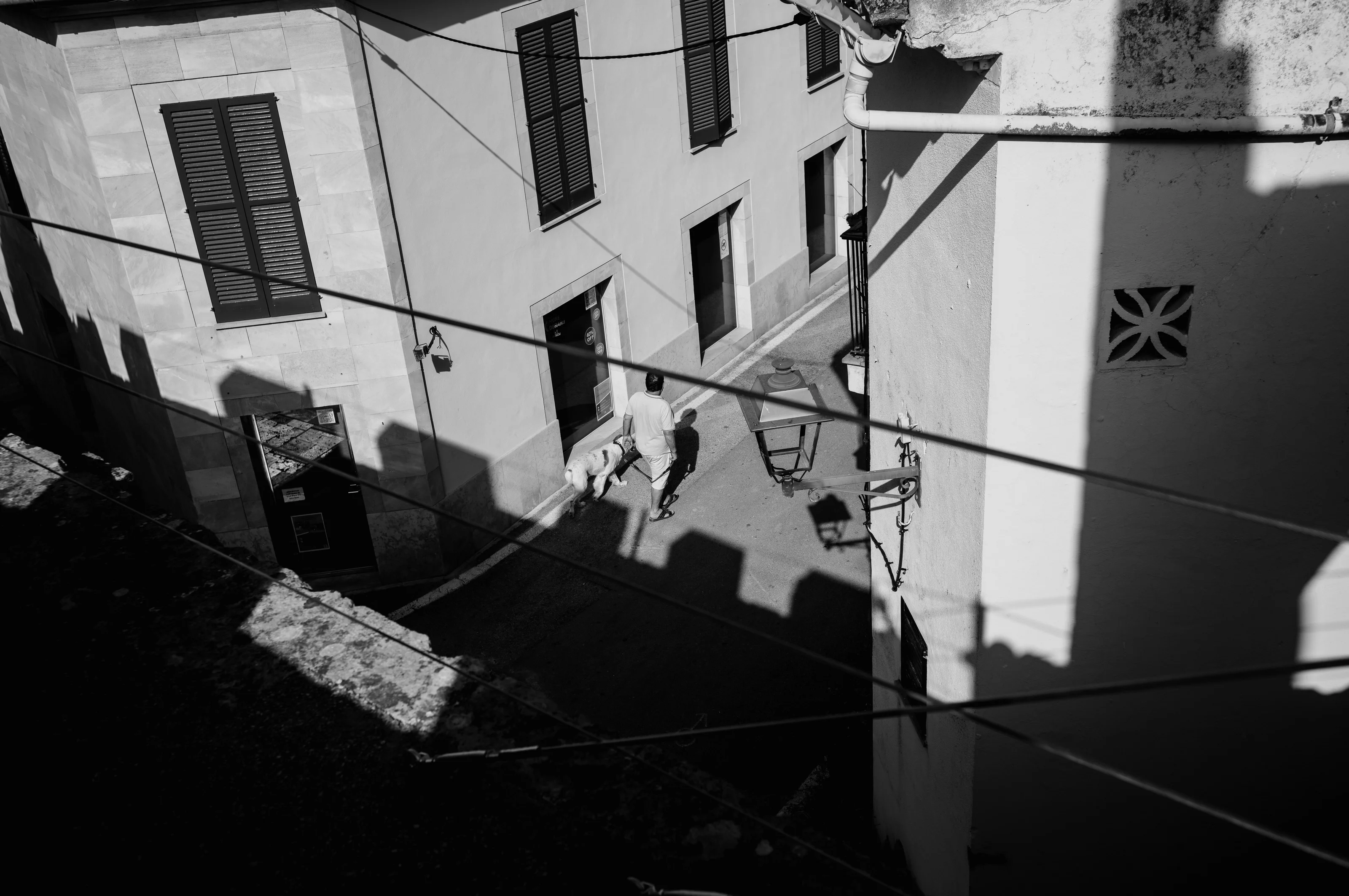Black and white photo of a person walking a dog in a narrow alley with overhead wires and shadows.