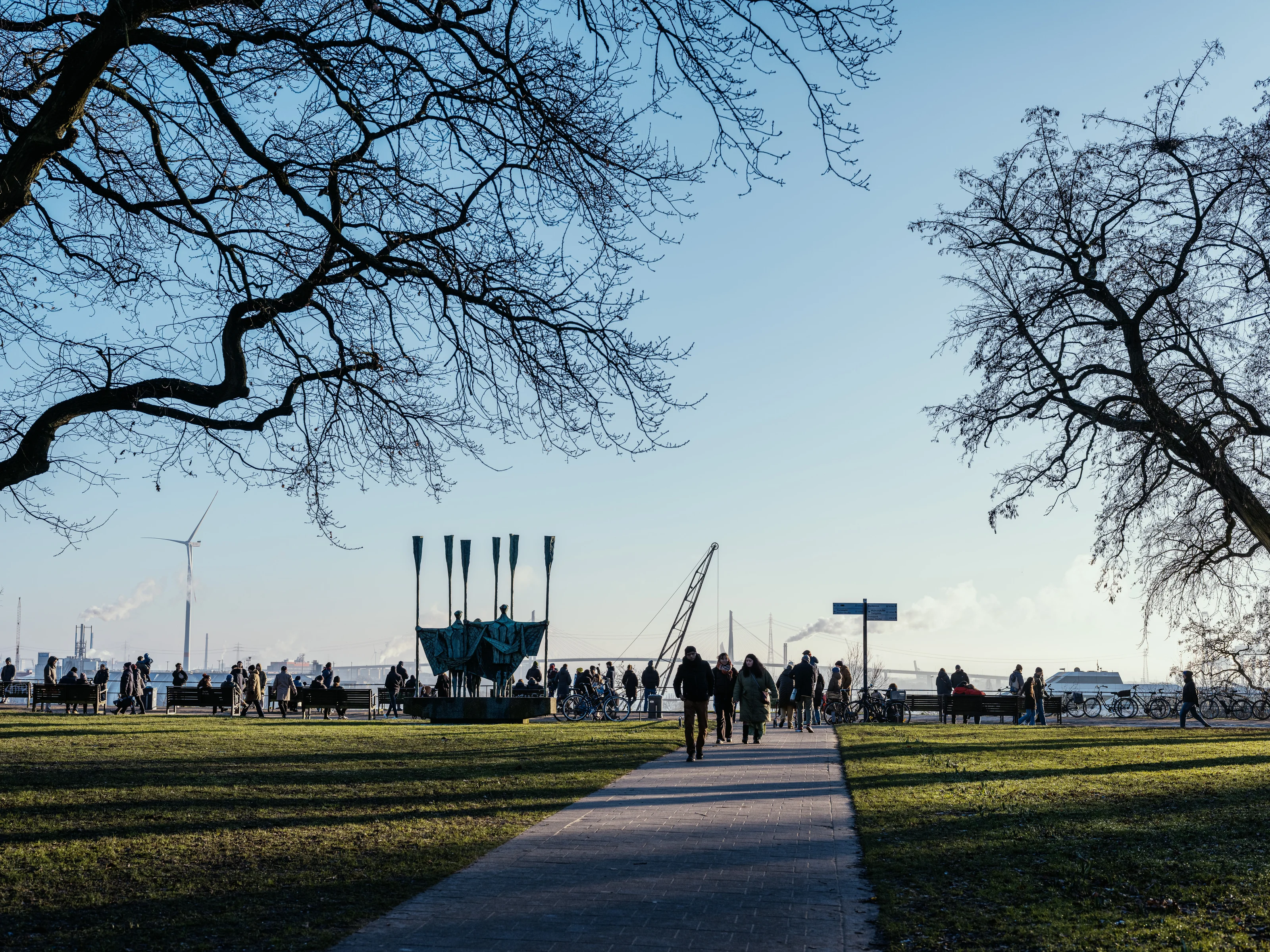 People walking in a park with leafless trees and an industrial backdrop.