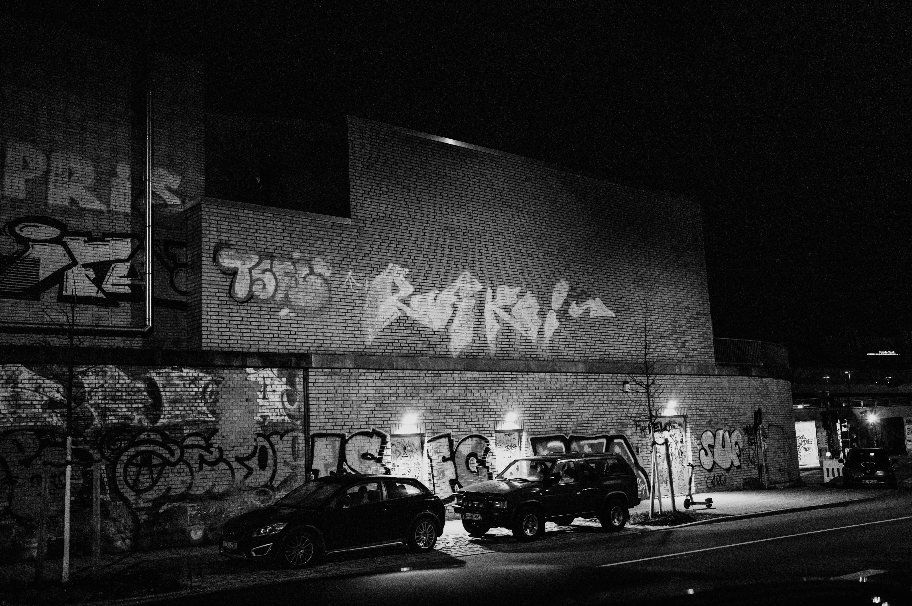 Black and white photo of a brick wall with graffiti and parked cars on a dimly lit street at night.
