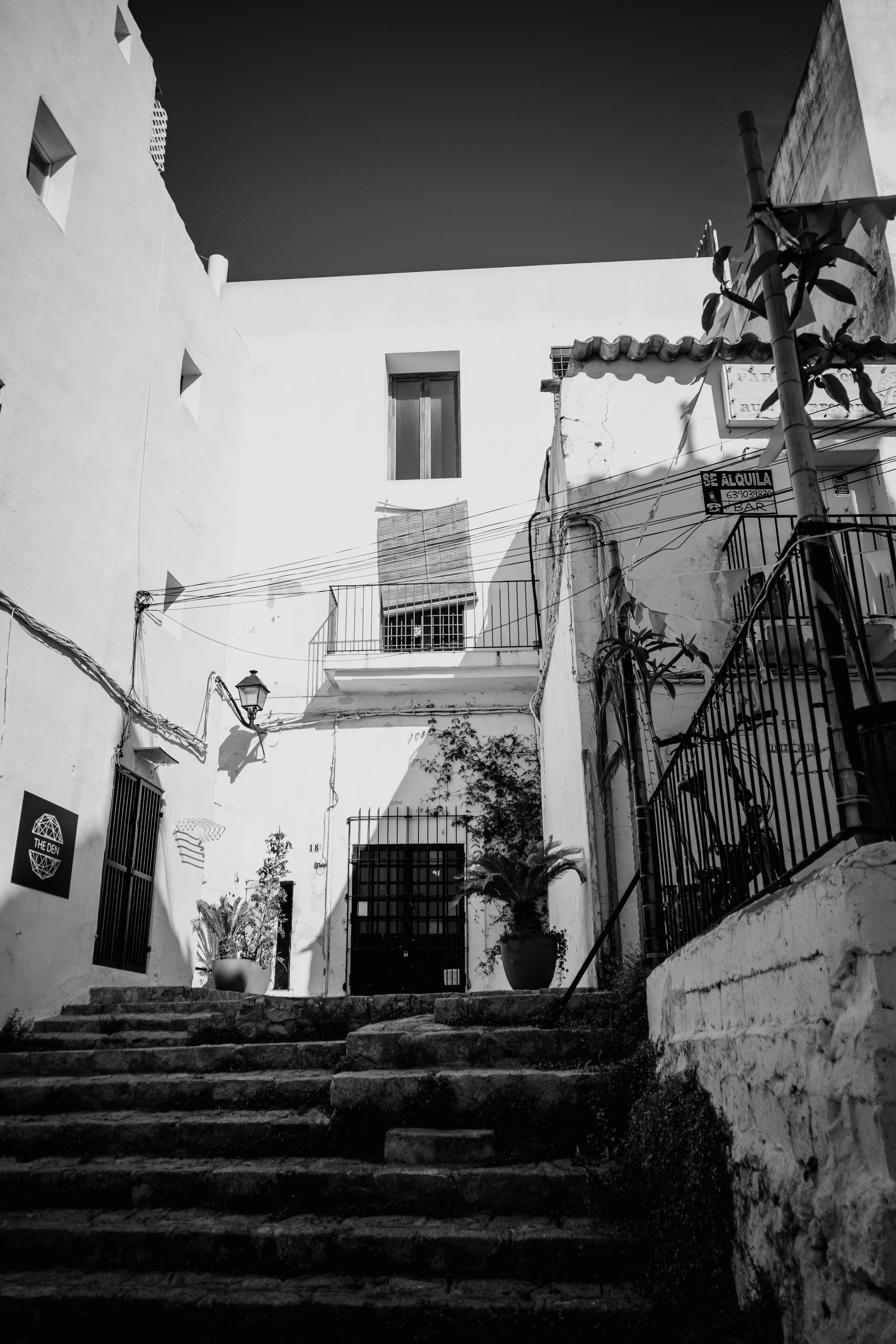 A black and white photo of a narrow alley with stone steps leading to a building with balconies and plants.