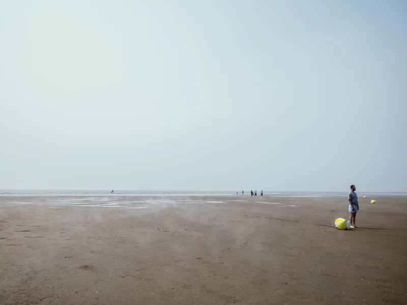 A person stands with a beach ball on a vast sandy beach with a hazy sky.