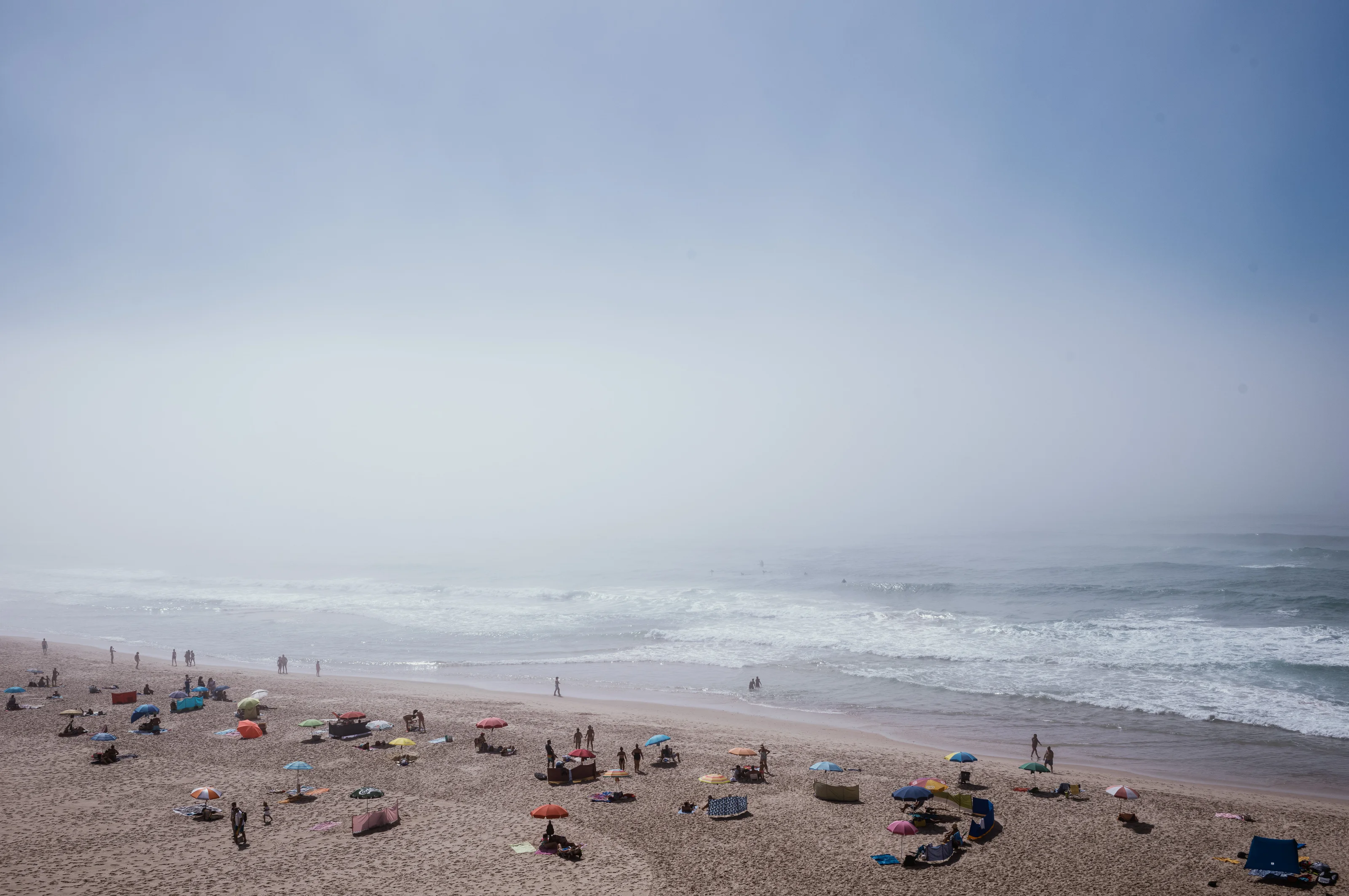 A foggy beach with several umbrellas and people scattered along the shore.
