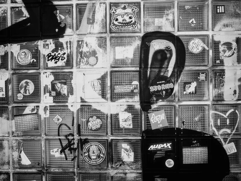 A monochrome wall with various stickers and graffiti art on glass blocks.
