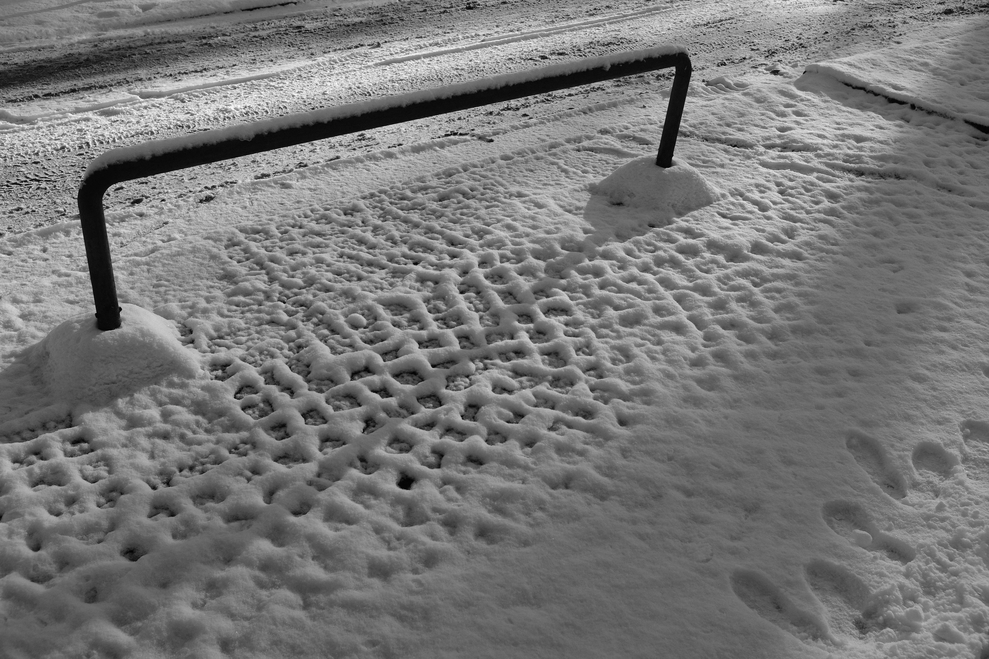 Snow-covered ground with tire and footprint impressions alongside a metal barrier.