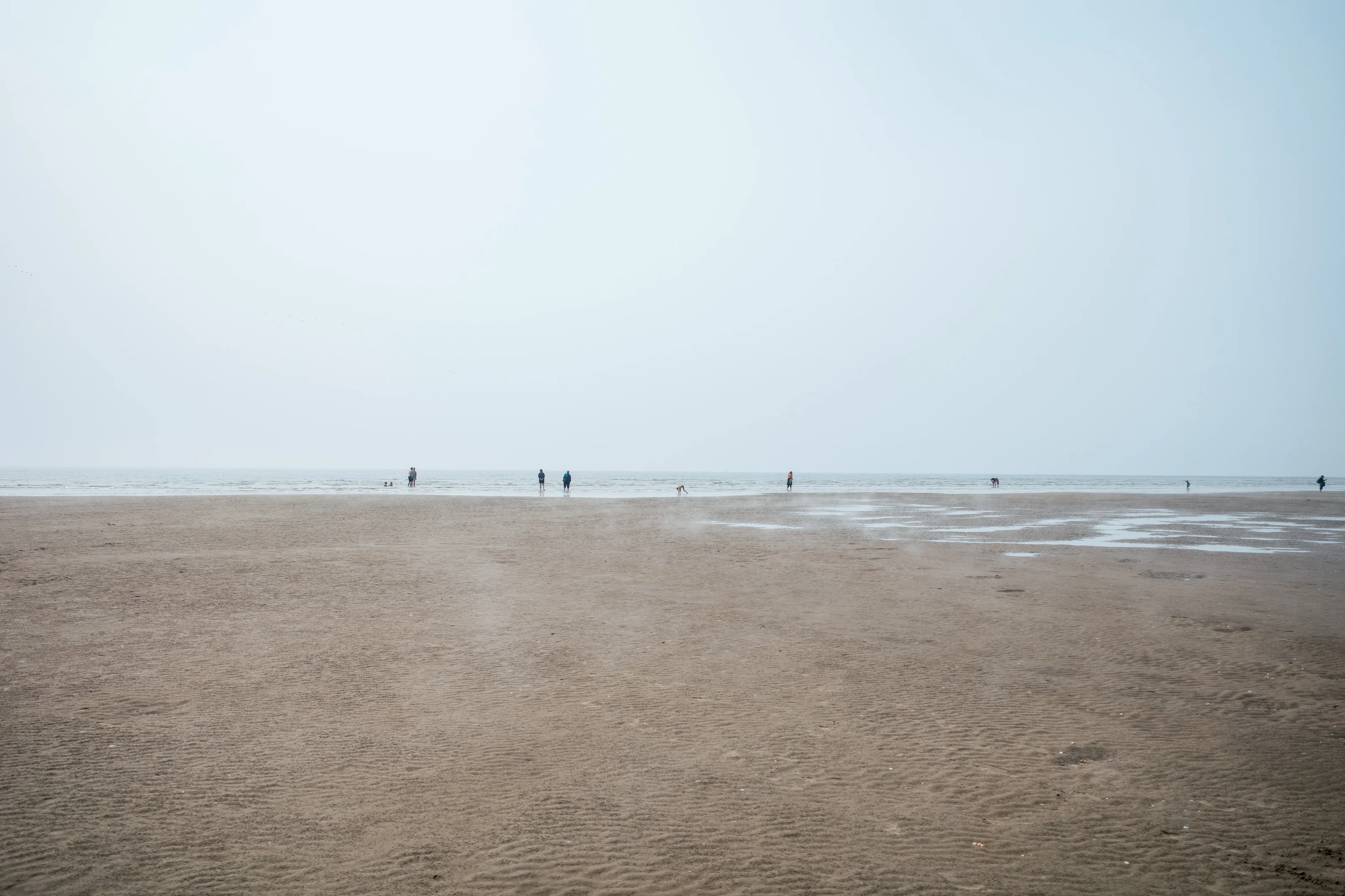 People walking on a wide, grayish beach under a hazy sky.