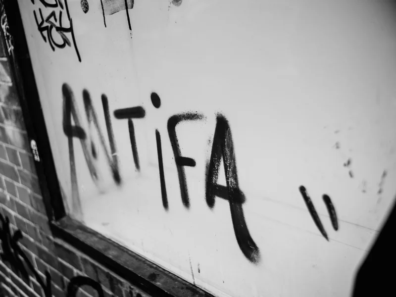 Black graffiti reading 'ANTIFA' on a white wall next to a brick surface.