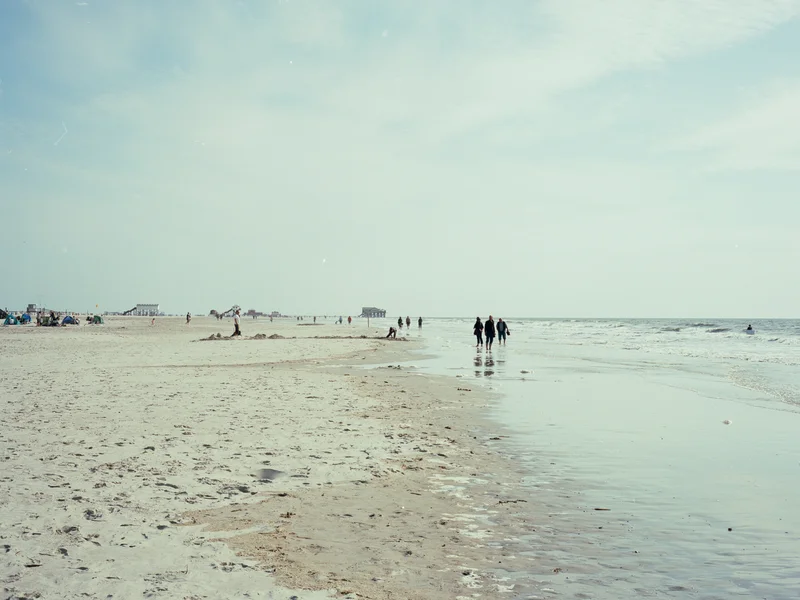 A wide sandy beach with people walking along the shoreline under a cloudy sky.