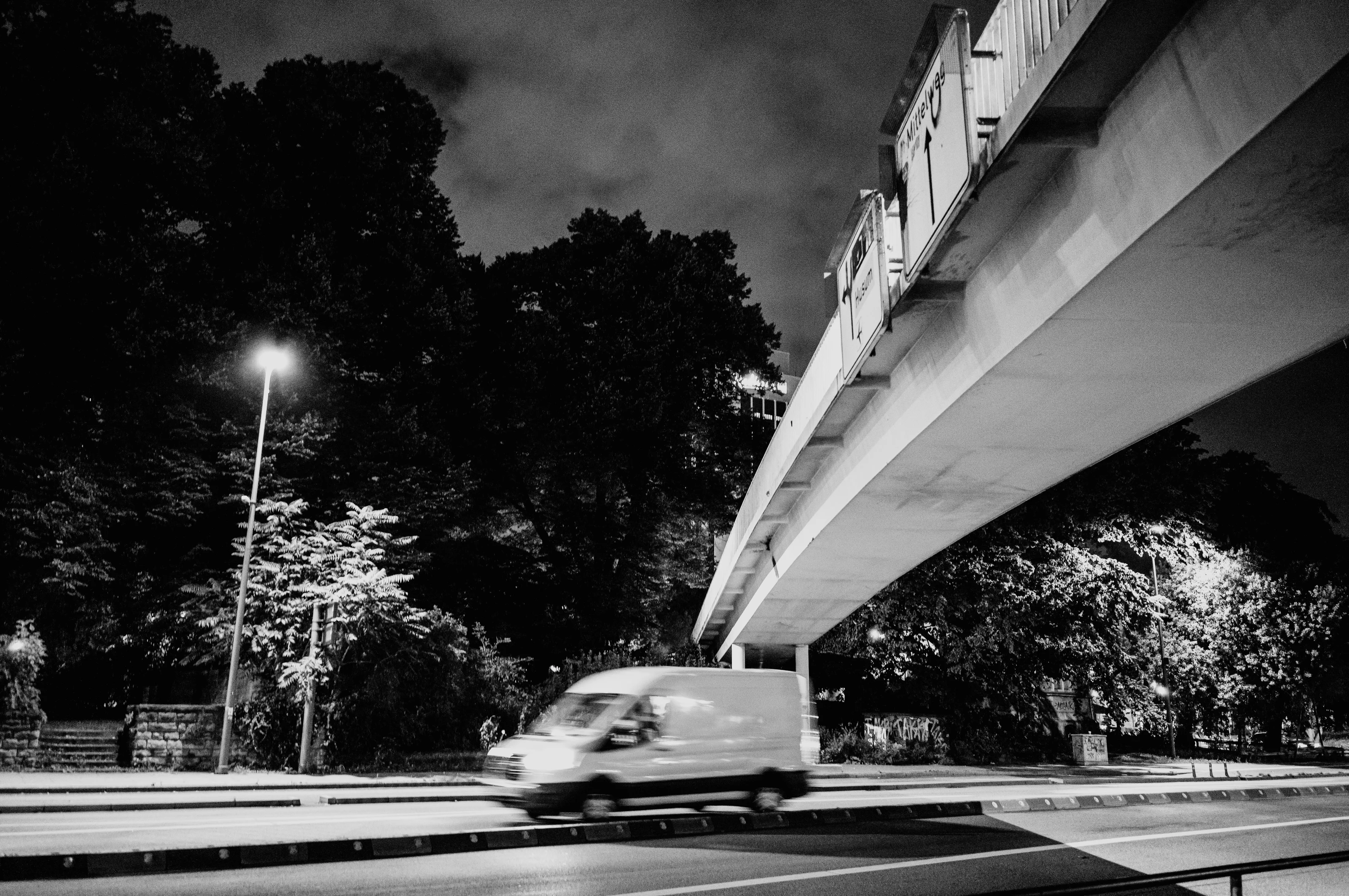 A van driving under a bridge at night with streetlights illuminating trees.