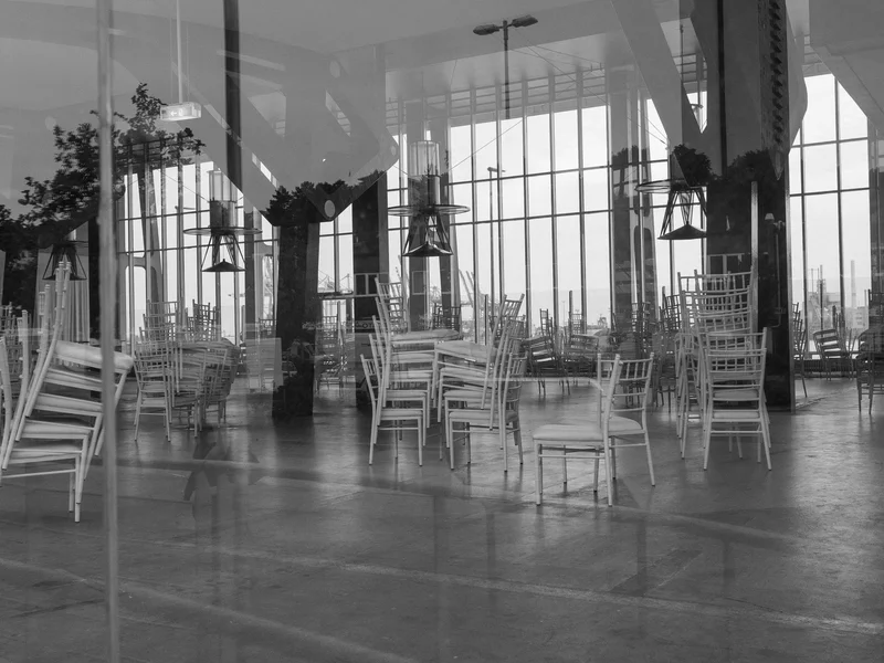 A black and white image of stacked chairs inside a room with large windows.
