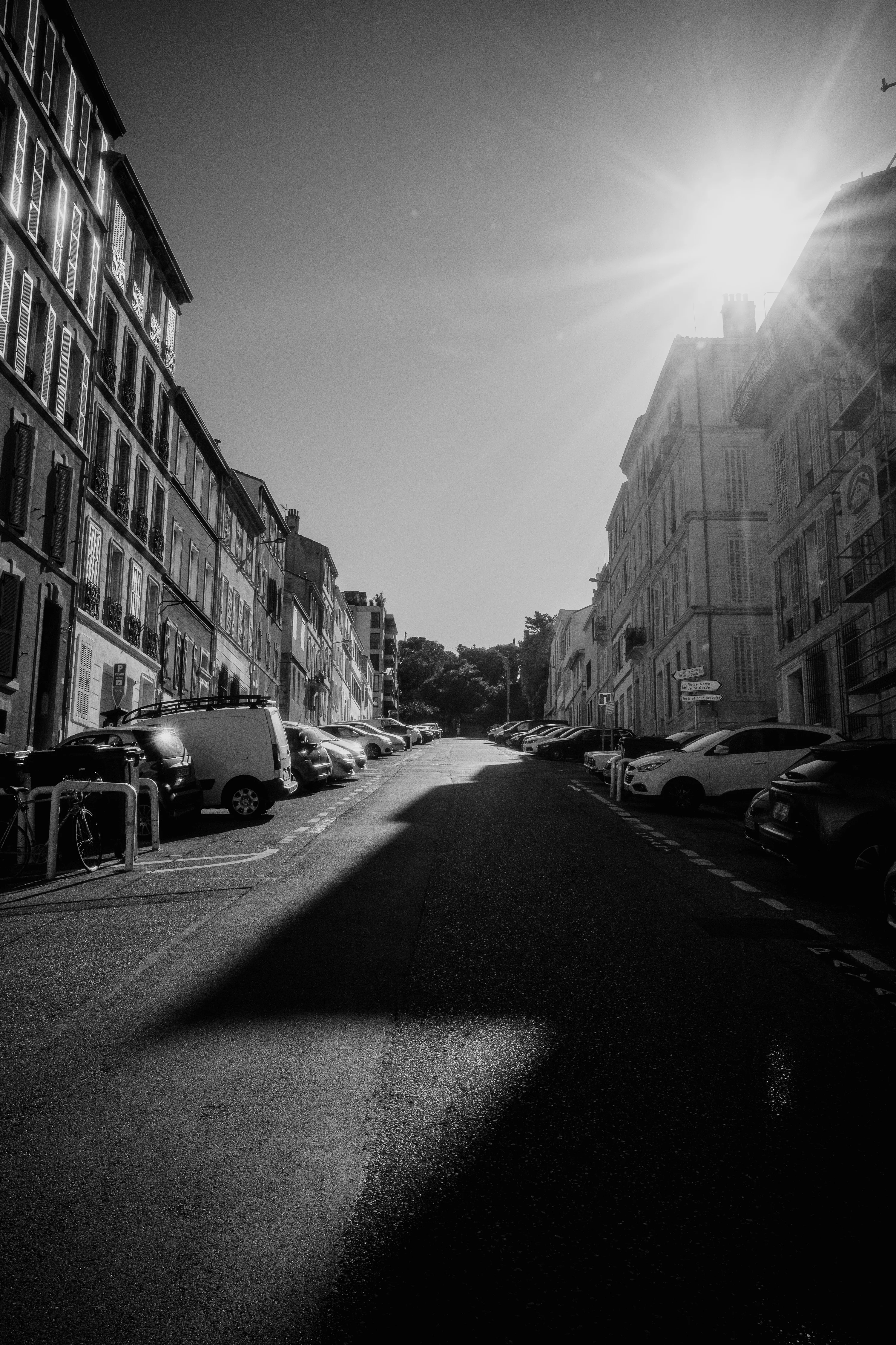 A sunlit street between rows of buildings with parked cars, casting long shadows.