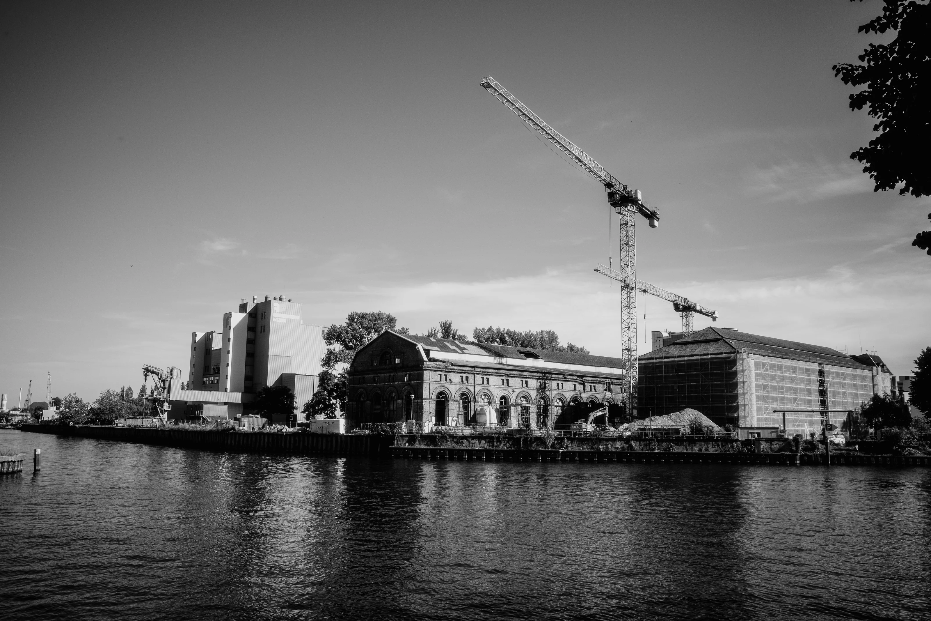 A black and white photograph of a riverside industrial site with cranes and large buildings.