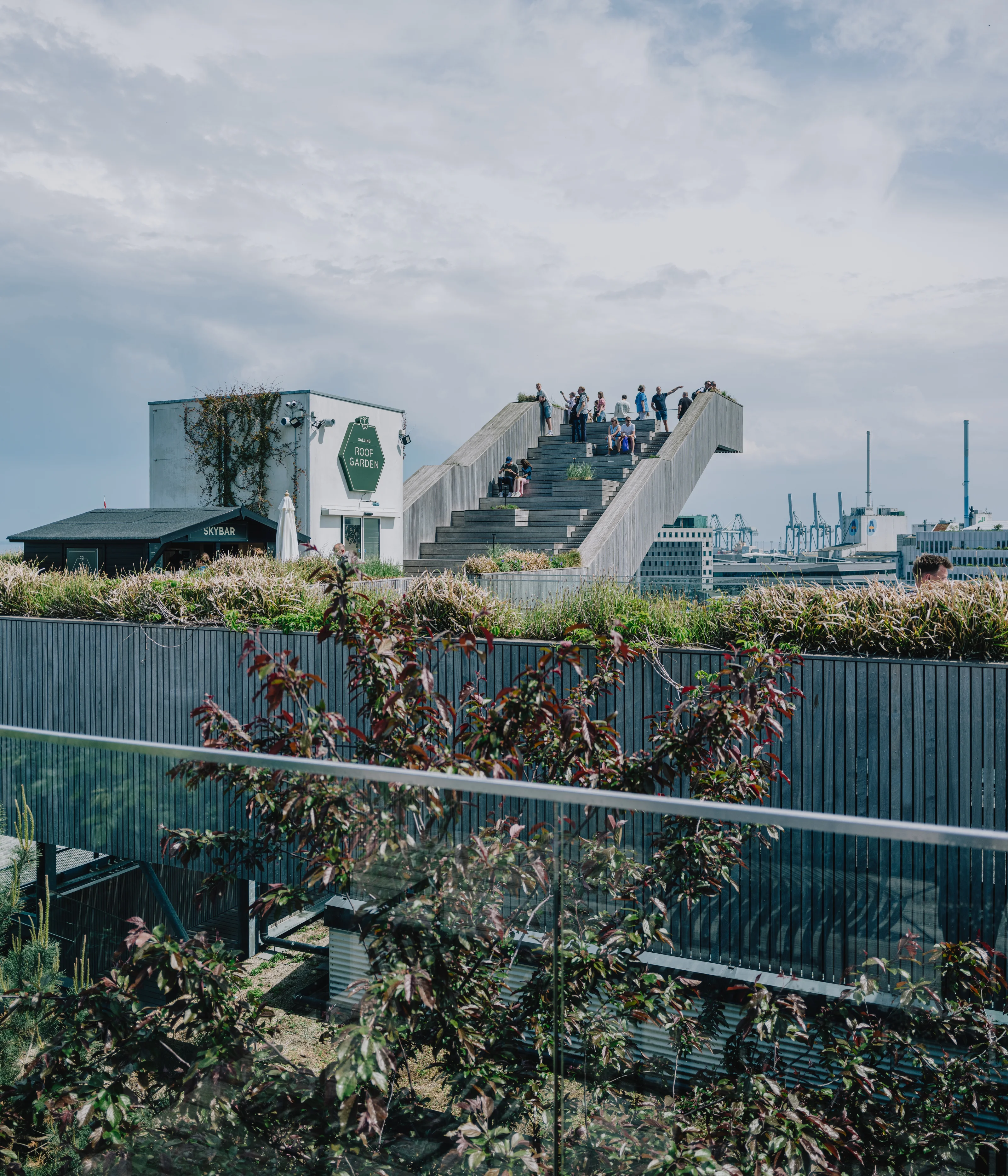 People standing on an elevated staircase structure surrounded by greenery and a cityscape in the background.
