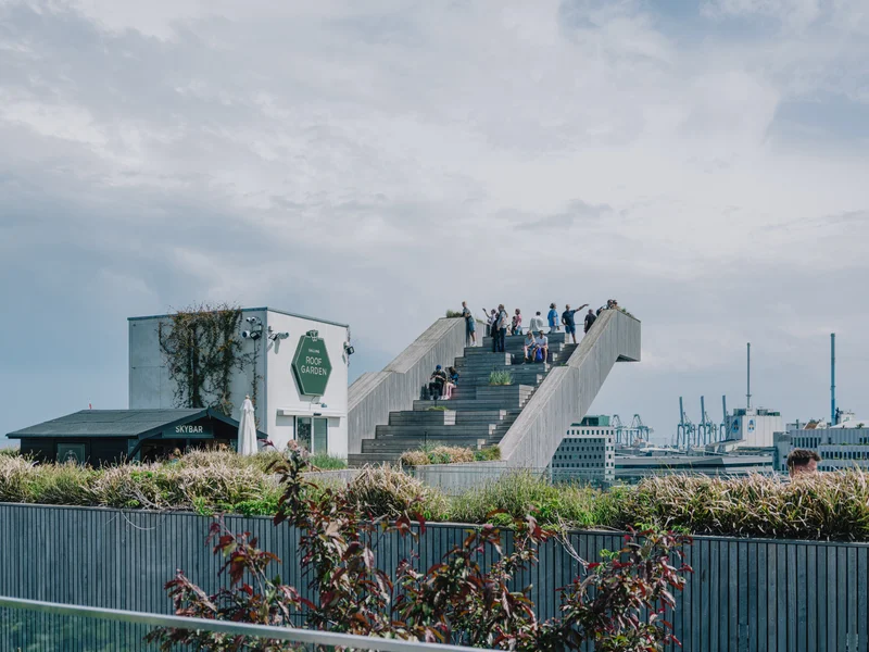 People standing on an elevated staircase structure surrounded by greenery and a cityscape in the background.