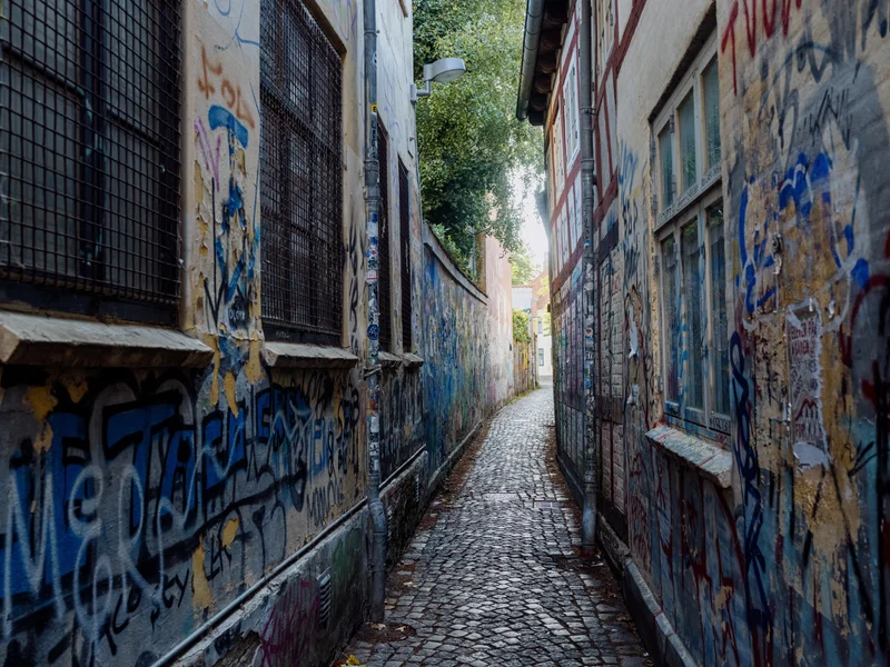 Narrow cobblestone alleyway with graffiti-covered walls and trees visible at the end.